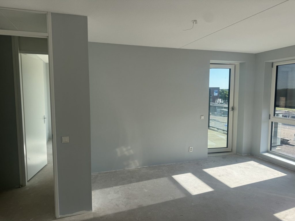 Nieuw appartement met kale, lichtgrijze wanden, een betonvloer en een balkondeur met uitzicht op een bouwterrein