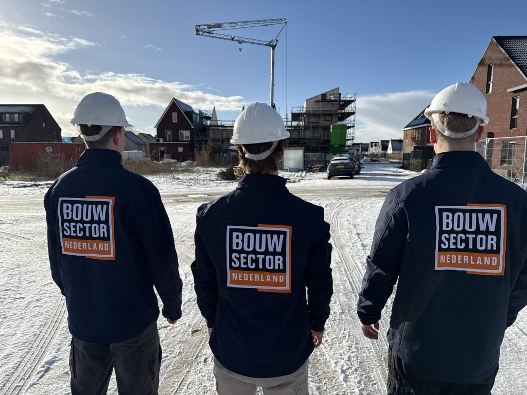 Drie vakmensen van Bouwsector Nederland met witte helmen en jassen met logo kijken naar nieuwbouwwoningen in aanbouw